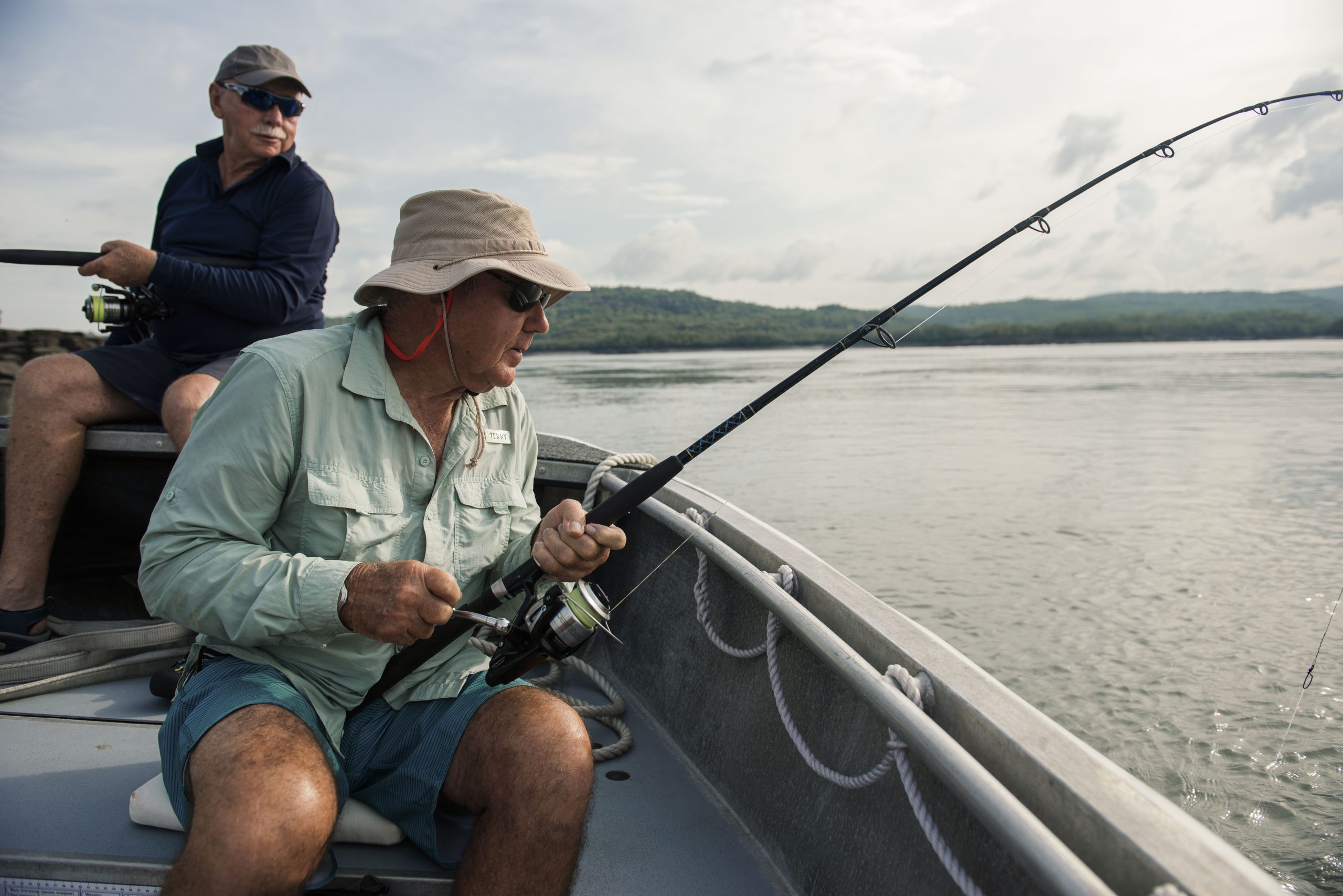 Two men fishing from an adventure boat in a calm Kimberley river, one actively reeling in a catch.