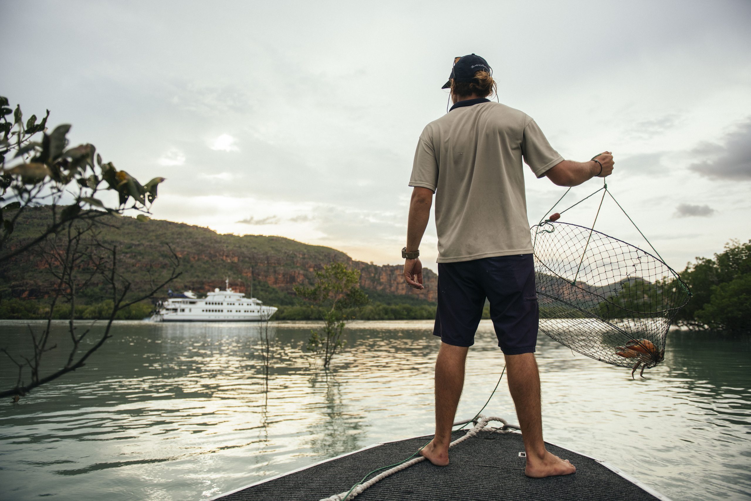 A True North guide standing on the edge of an adventure boat holding a crab pot with a mud crab, with TRUE NORTH cruise ship anchored in the background among Kimberley cliffs.
