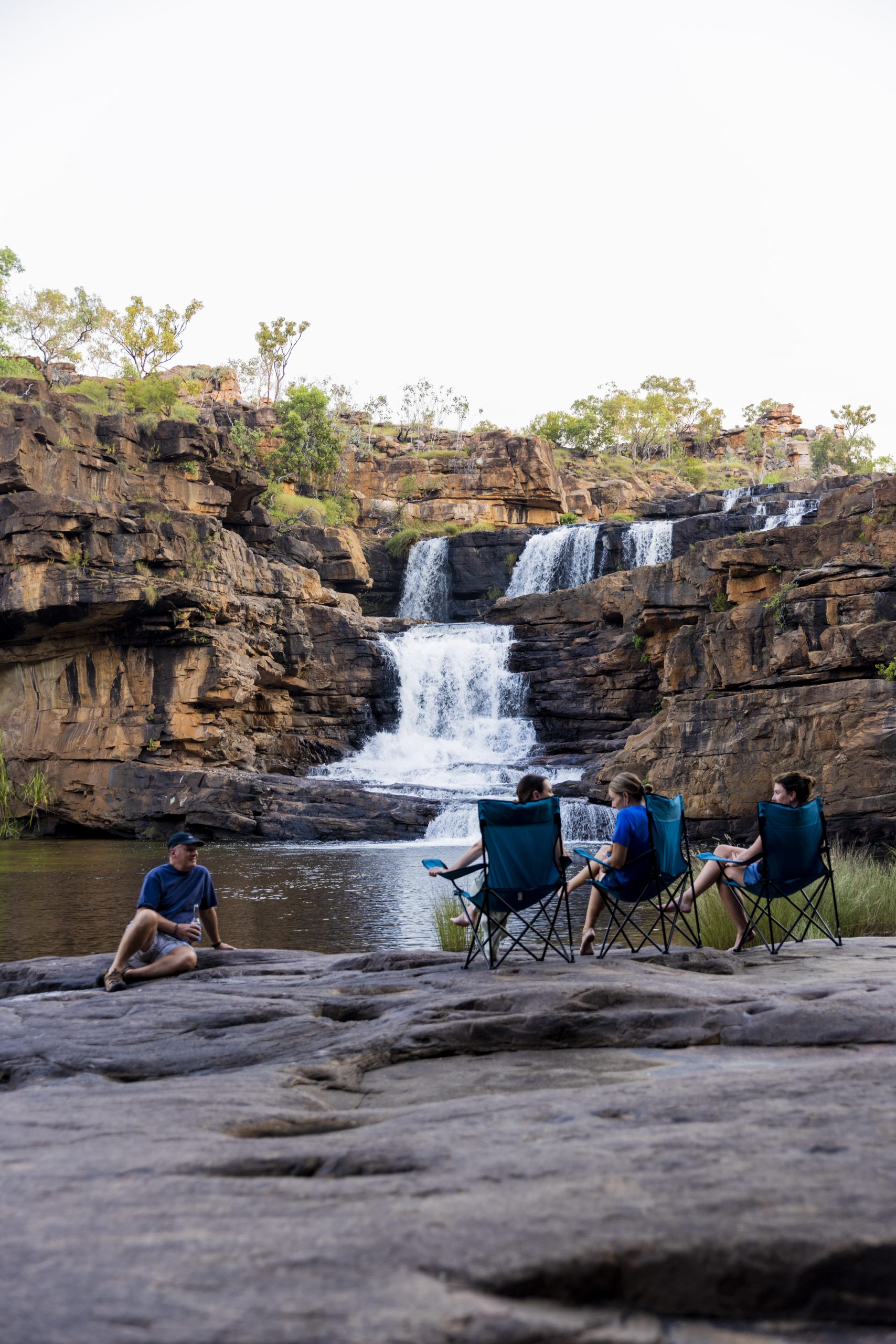 Guests relaxing in camp chairs beside Eagle Falls during a remote heli-picnic with True North in the Kimberley, surrounded by rugged sandstone cliffs and cascading water